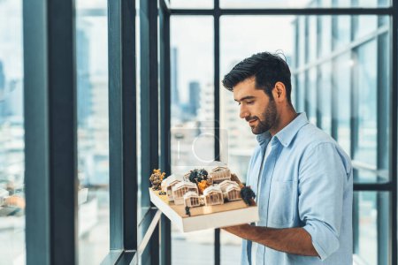 Photo for Businessman in casual outfit holding house model while checking house construction. Architect engineer inspect building model while standing near window with skyscraper. Civil engineering. Tracery. - Royalty Free Image
