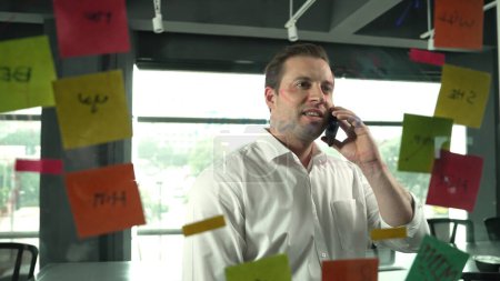 Photo for Professional businessman making phone call to customers while looking at marketing strategy idea written on colorful sticky notes. Project manager talking on phone analysis data on wall. Manipulator. - Royalty Free Image