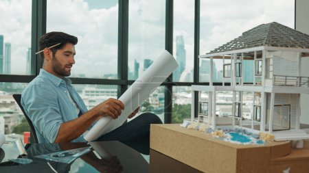 Photo for Smiling engineer looking project plan while design house model at office. Smart caucasian sitting relaxing while reading project plan. Creative design, civil engineering. Skyscraper. Tracery - Royalty Free Image