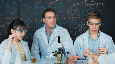 Kreativer Lehrer zeigt Chemie auf Tafel, während er mit einem Gymnasiasten am Tisch mit Mikroskop und Reagenzglas mit farbiger Flüssigkeit spricht. Junge Studentin im Laborkittel im MINT-Unterricht. Erbauung.
