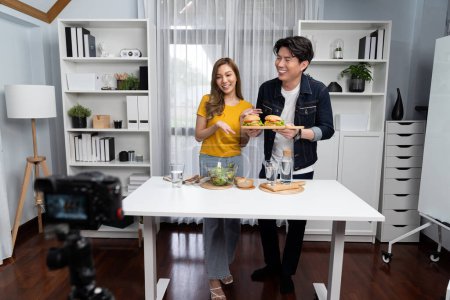 Photo for Chef influencers presenting on cooking show on social media channel serving double big hamburger set with bread and salad menu side dish on live streaming recording by video camera at home. Infobahn. - Royalty Free Image