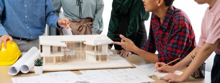 Photo for Architect team analysis and brainstorming about house construction at meeting table with house model, blueprint and architectural equipment scatter around. Creative design and teamwork. Burgeoning. - Royalty Free Image