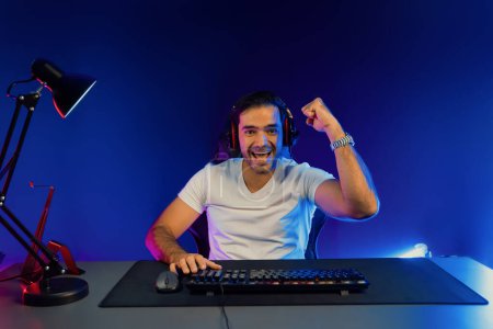 Photo for Winner of smart streamer playing online game wearing headphones raising fist up on streaming gamer with social media. Esport skill team player online digital gaming at neon light studio room. Surmise. - Royalty Free Image
