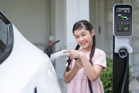 Photo for Happy little young girl learn about eco-friendly and energy sustainability as she recharge electric vehicle from home EV charging station. EV car and sustainable future generation concept. Synchronos - Royalty Free Image
