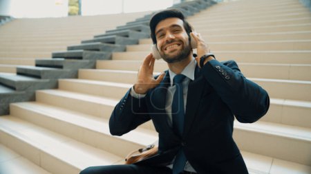 Photo for Project manager listening and enjoy music from headphone while sitting at stairs. Professional businessman wearing suit outfit while dancing and moving to music. Happy man listen funny song. Exultant. - Royalty Free Image