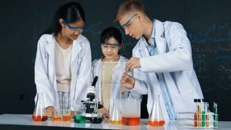 Photo for Smart girl pour colored solution in to beaker while diverse student excited about doing experiment. Professional scientist prepare for doing experiment at blackboard with chemical theory. Edification. - Royalty Free Image