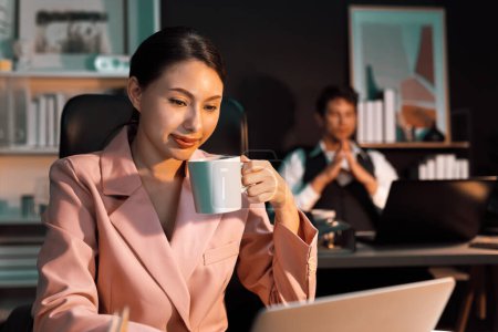 Photo for Focusing on sitting woman coworker drinking tasty coffee on working front desk while waiting email business project report at late night time with blurry man background working on laptop. Postulate. - Royalty Free Image
