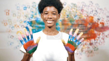 Photo for African boy looking at camera while showing two side of hand to camera with colorful color. Smiling happy highschool teenager wearing white shirt while standing at colorful stained wall. Edification. - Royalty Free Image