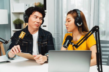 Photo for Smiling radio influencer hosts wearing headphones with channel social media online live streaming with talking topic on script note and laptop to listeners at morning time at studio record. Infobahn. - Royalty Free Image