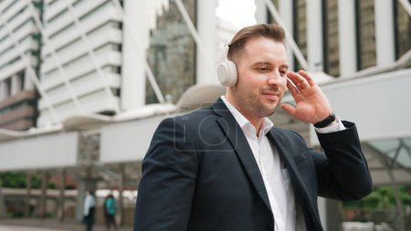 Glücklicher Geschäftsmann, der über Kopfhörer Entspannungsmusik hört und sich mit Musik bewegt, während er mit lebhafter Stimmung auf der Straße in der Großstadt spaziert. Manager tragen Headset und hören entspannt Reimen zu. Urbane Pflanzen.