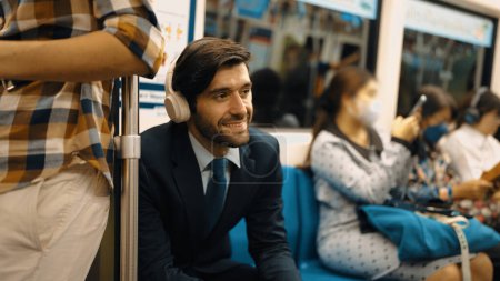 Téléchargez les photos : Un bel investisseur souriant écoutant de la musique relaxante tout en étant assis dans le train. Homme d'affaires professionnel aiment écouter de la musique tout en utilisant les transports publics au travail. Fond flou. Exultant. - en image libre de droit