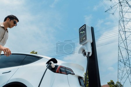 Photo for Man recharge EV electric car battery at charging station connected to electrical power grid tower on sky background as electrical industry for eco friendly vehicle utilization. Expedient - Royalty Free Image