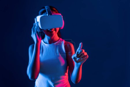 Foto de Soporte femenino inteligente con auriculares VR rodeados de luz de neón cyberpunk que conectan tecnología de comunidad ciberespacial metaversa y futurista. Mujer usando el dedo señalando objeto virtual. Alucinaciones. - Imagen libre de derechos