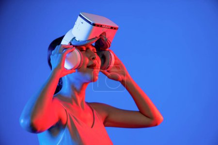 Téléchargez les photos : Smart femelle stand frappé par la lumière néon cerclage casque VR au-dessus de l'œil.La fonction gadget pour connecter métaverse, future technologie cyberespace. Femme lever les bras tenant des lunettes à la main. Hallucination. - en image libre de droit