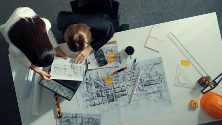 Photo for Top view of businesswoman hand blueprint to engineer sitting at meeting table with project plan, equipment and safety helmet. Aerial view of manager explain about building construction. Alimentation. - Royalty Free Image
