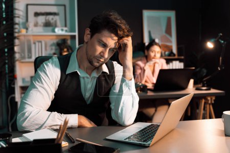 Photo for Sitting man with serious face thinking creative business project planning at night lighting time at modern office with blurry secretary checking paper report with folder background concept. Postulate. - Royalty Free Image