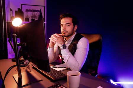 Foto de Hombre de negocios inteligente trabajando remotamente en el escritorio con el mercado objetivo de investigación en el canal de ventas de la base de datos de productos de servicio en línea en la pantalla de PC en la oficina de casa de luz oscura de neón púrpura a altas horas de la noche. Conjetura. - Imagen libre de derechos