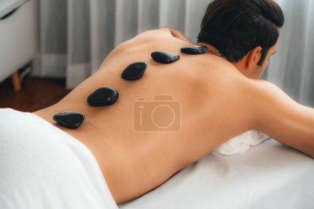 Téléchargez les photos : Massage aux pierres chaudes au salon de spa dans un complexe de luxe avec lumière du jour sérénité ambiante, homme heureux client profitant d'un massage aux pierres de basalte spa glisser sur le corps avec une chaleur apaisante. Tranquillité - en image libre de droit