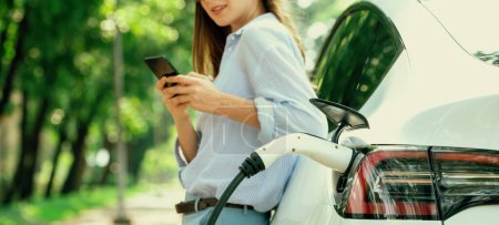 Téléchargez les photos : Jeune femme utilisant smartphone application bancaire en ligne pour payer la charge de la batterie de voiture électrique à partir de la station de charge EV pendant le voyage de vacances sur la route dans le parc national ou la forêt d'été. Panorama Exalt - en image libre de droit