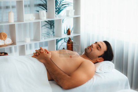 Téléchargez les photos : Homme caucasien client appréciant relaxant massage spa anti-stress et dorloter avec beauté loisirs de la peau dans la lumière du jour salon de spa d'ambiance dans un complexe de luxe ou un hôtel. Tranquillité - en image libre de droit