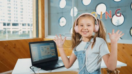 Chica sonriente mirando mientras agita la mano a la cámara con el ordenador portátil colocado en la mesa. Niño que usa auriculares sonriendo mientras la pantalla del portátil muestra el programa de programación o codificación del sistema en la clase STEM. Erudición.