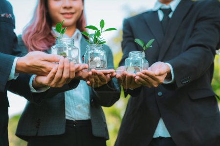 Concept of sustainable money growth investment with glass jar filled with money savings coins with businesspeople as eco-friendly financial investment nurtured with nature and healthy retirement. Gyre