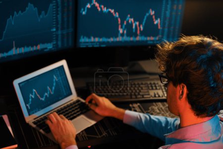 Foto de Joven inversor stock trader que busca en el ordenador portátil en comparación con las pantallas dinámicas de tipo de cambio del gráfico de mercado actual en línea en tiempo real en neón luz oficina moderna, el análisis de datos financieros. Gusher.. - Imagen libre de derechos