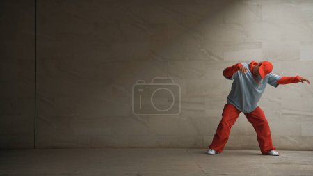Foto de Guapo coreógrafo practicar danza callejera en fondo gris. Joven y atractiva bailarina b boy realizar mientras usa un paño elegante. Sensación de paso animado. Copiar espacio. Deporte al aire libre 2024. Rígidamente.. - Imagen libre de derechos
