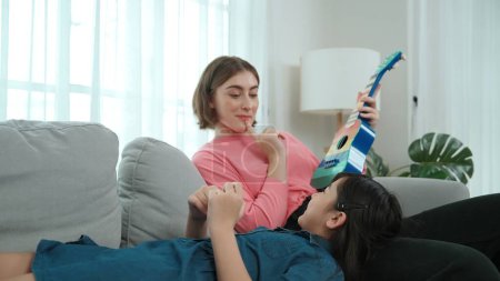 Asian cute daughter listening acoustic music while lie on her mother lap at sofa. Caucasian mother playing colorful ukulele while cute child looking at her parent. Family recreation concept. Pedagogy.