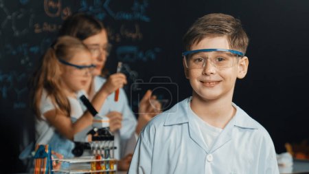 Photo for Smart boy crossing arms while teacher and friend doing experiment at STEM science class. Student mixing colored solution while standing at board written chemistry theory at laboratory. Erudition. - Royalty Free Image