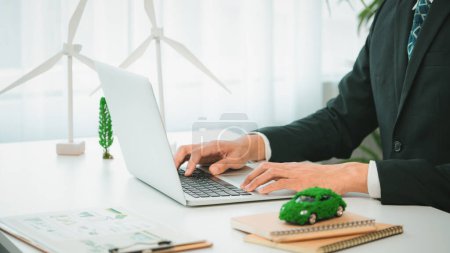 Businessman working in office developing plan or business project on eco-friendly ev car using net zero alternative energy technology on computer for greener environment as apart of CSR effort. Gyre