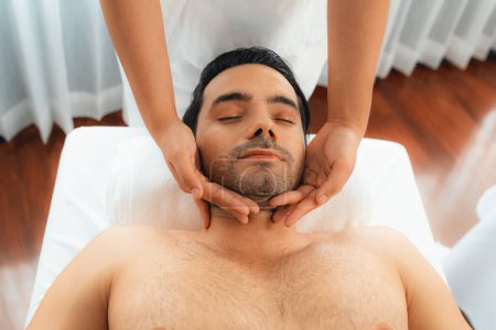 Téléchargez les photos : Homme caucasien appréciant relaxant massage de la tête anti-stress et dorloter beauté faciale loisirs de la peau dans dayspa lumière moderne ambiant au complexe de luxe ou salon de spa de l'hôtel. Tranquillité - en image libre de droit