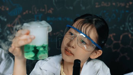 Junges Mädchen hält grüne chemische Lösung in der Hand und inspiziert sie sorgfältig, während sie im Labor vor der Tafel mit biochemischer Theorie sitzt. Nahaufnahme einer jungen Studentin beim Experimentieren. Erbauung.