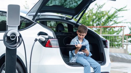 Photo for Little boy sitting on car trunk, using tablet while recharging eco-friendly car from EV charging station. EV car road trip travel as alternative vehicle using sustainable energy concept. Perpetual - Royalty Free Image