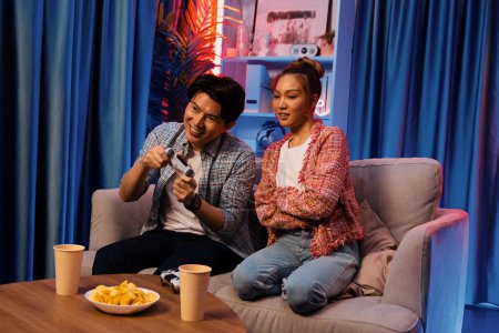 Photo for Sitting sofa of happy couple playing video game with joysticks, smiling man playing challenge, raising fist up to be winner the challenge at modern neon blub light living room at home. Infobahn. - Royalty Free Image