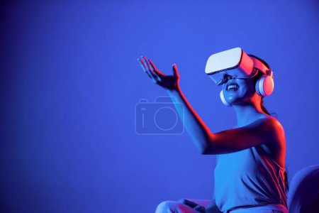 Photo for Smart female sitting on sofa surrounded by neon light wear VR headset connect metaverse, futuristic cyberspace community technology, watch and interacting with virtual object with hand. Hallucination. - Royalty Free Image