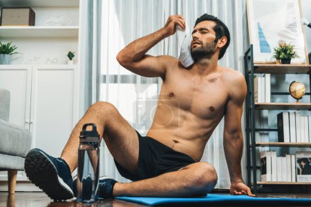 Photo for Athletic and sporty man sitting, resting on fitness mat after finishing home body workout exercise session for fit physique and healthy sport lifestyle at home. Gaiety home exercise workout training. - Royalty Free Image