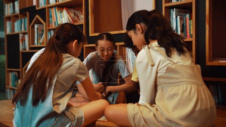 Photo for Group of diverse children sitting in circle while reading a book at library. Highschool girl sharing a magazine while pointing at interested topic and talking with multicultural friends. Edification. - Royalty Free Image