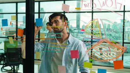Photo for Professional businessman uses sticky notes at glass wall with graph to brainstorming idea while multicultural businesspeople discussing about marketing ideas at office with city view. Tracery - Royalty Free Image