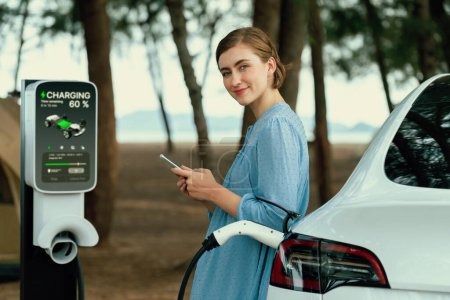 Photo for Holiday road trip vacation traveling to the beach camp with electric car, young woman checking battery from smartphone while recharge EV vehicle. Beach travel camping with eco-friendly car .Perpetual - Royalty Free Image