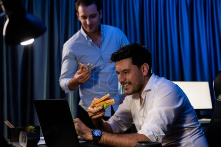 Foto de Compañeros de trabajo con cara feliz comiendo sándwich y tomando café mientras trabajan en el proyecto de trabajo por la noche. Freelancers creativos que reciben la aplicación de red en línea ganadora de ofertas por correo electrónico. Vendible. - Imagen libre de derechos