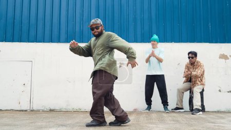 Asiatischer Hipster beim Salto vor der Wand mit einem lebhaften Freund. Attraktive Straßentänzerin übt Breakdance auf der Straße mit blauem Hintergrund, umgeben von Freunden. Outdoor-Sport 2024. Vorfreude.