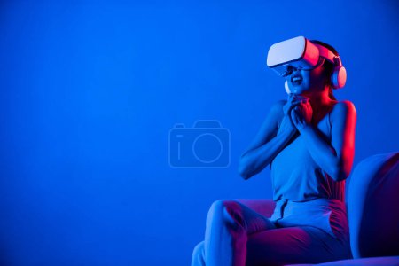 Photo for Smart female sitting on sofa surrounded by neon light wear VR headset connect metaverse, futuristic cyberspace community technology. Elegant woman excited and emotionally watch movie. Hallucination. - Royalty Free Image
