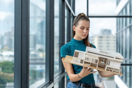 Photo for Beautiful young architect engineer holds architectural model while inspect house model. Professional interior designer checking house construction while standing near window with city view. Tracery. - Royalty Free Image