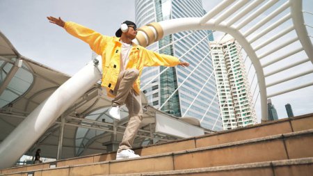 Foto de Elegante asiático hombre realizar hiphop bailando con bajo ángulo de la cámara. Bailarina de descanso subiendo la escalera mientras escuchan música de auriculares y bailan juntos en la ciudad. Deporte al aire libre 2024. Rígidamente.. - Imagen libre de derechos