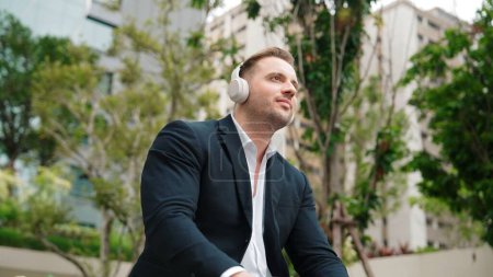 Photo for Professional businessman wearing headphone and while moving to relaxing music in green city. Caucasian project manager dancing to song while sitting and relaxed at eco urban city. Lifestyle. Urbane. - Royalty Free Image