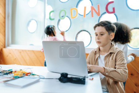 Photo for The schoolboy in classroom sitting and learning about engineering remote control with laptop while other child watch outside. Circuit wire, motherboard and notebook put on desk messily. Erudition. - Royalty Free Image