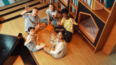 Photo for Top view of diverse children sitting in circle while looking at camera and waving hands at library surrounded with the stack of book. Multicultural highschool student greeting camera. Edification. - Royalty Free Image