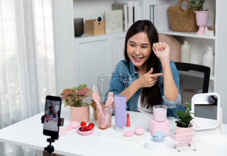 Joven hermosa asiática mostrando maquillaje cosmético lápiz labial mate pastel prueba de color en la colección bestseller en brazo prueba promoción en las redes sociales grabación en línea smartphone en la habitación moderna. Estratagema.
