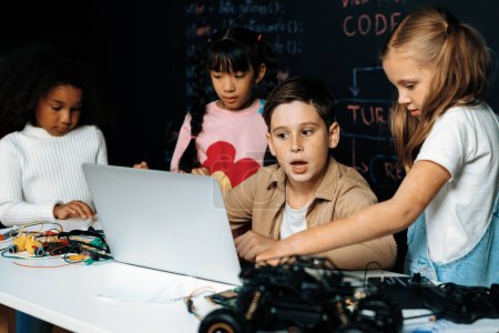 Photo for In classroom student in STEM class learning about coding robotics car. Brown shirt Schoolboy and white bib schoolgirl watching laptop and discussing. Other schoolgirls playing around funny. Erudition. - Royalty Free Image